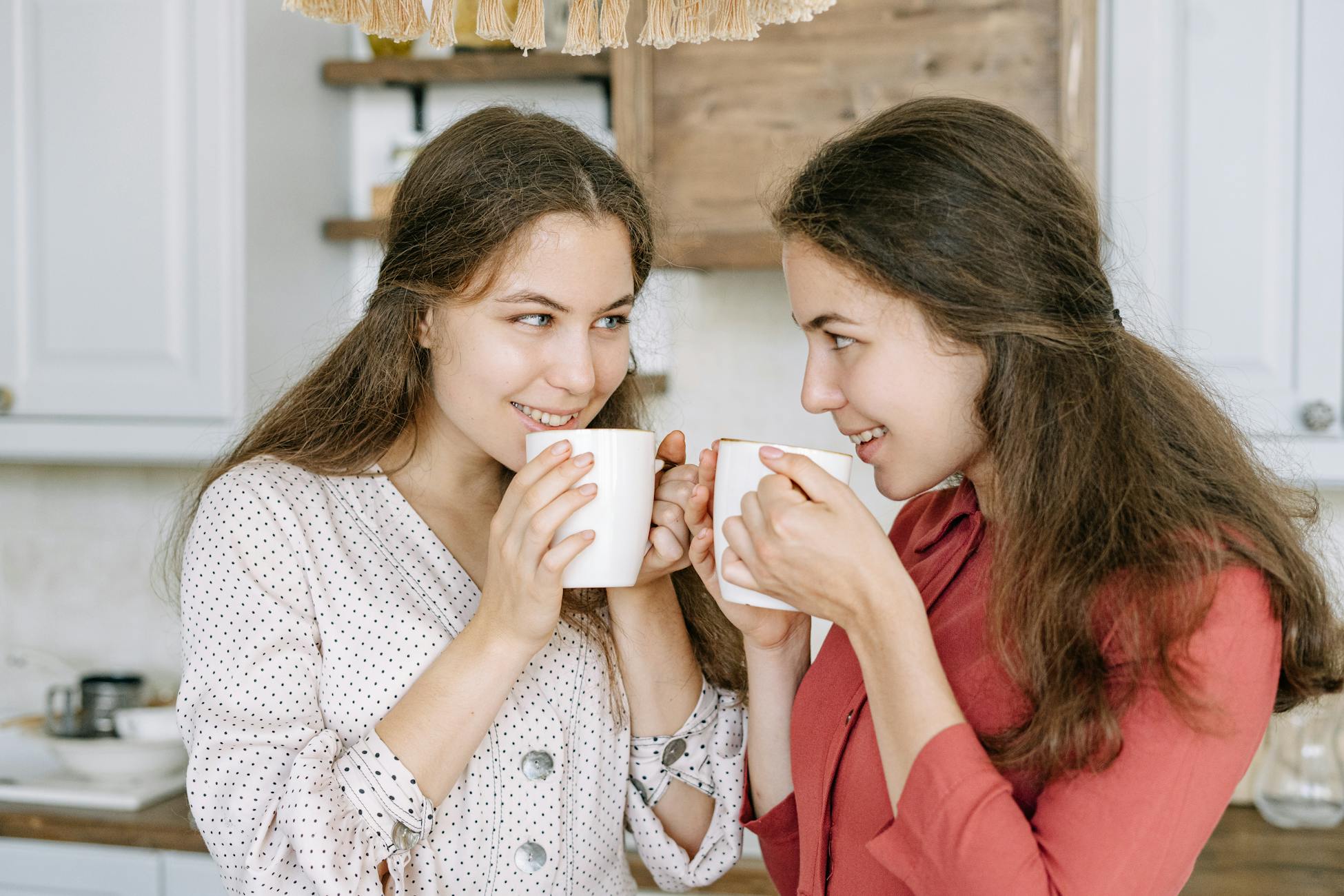Two women smiling and enjoying hot beverages indoors, creating a warm, cozy atmosphere.