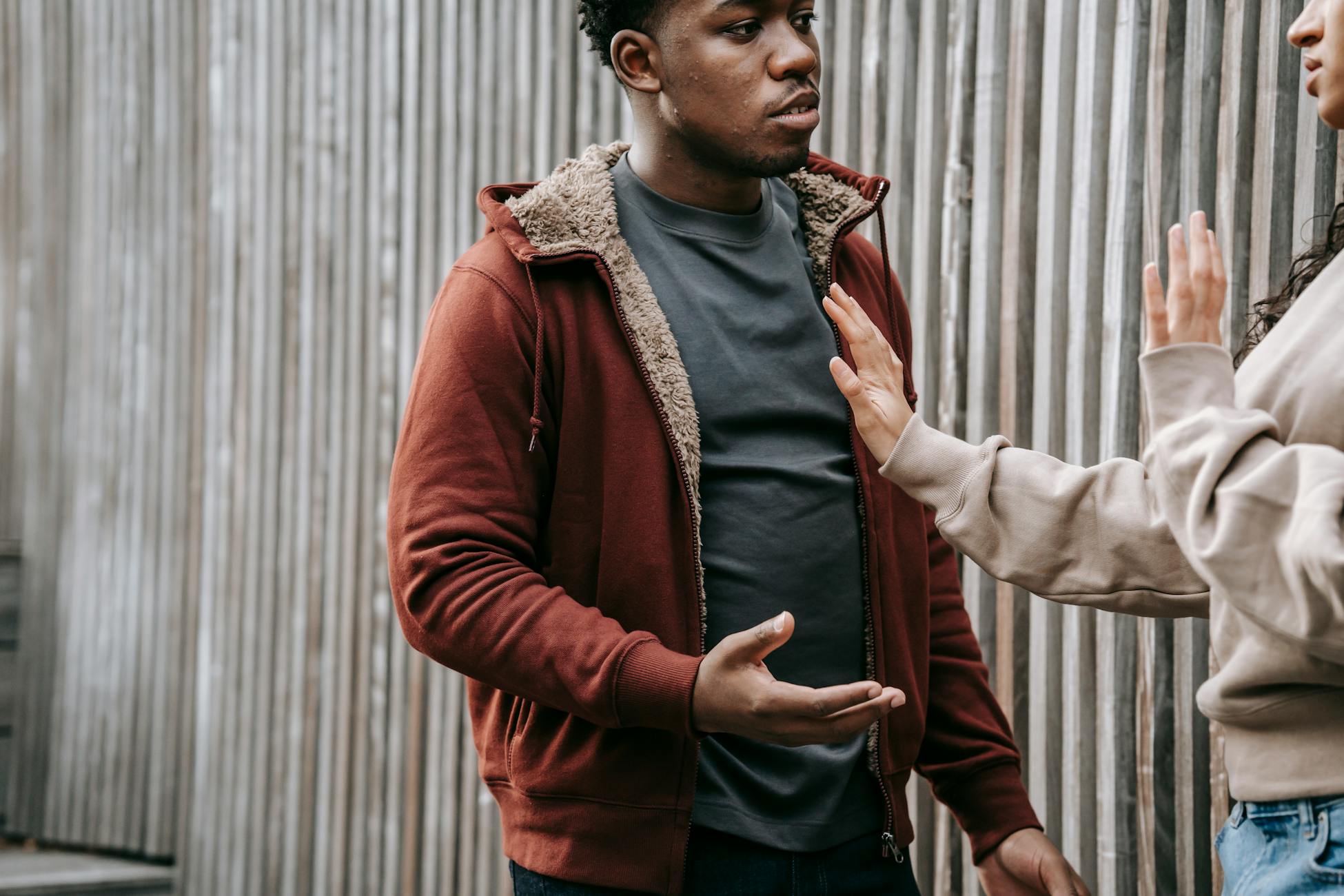 Unrecognizable woman with outstretched arm trying to stop irritated African American man while having argument on street near wooden fence