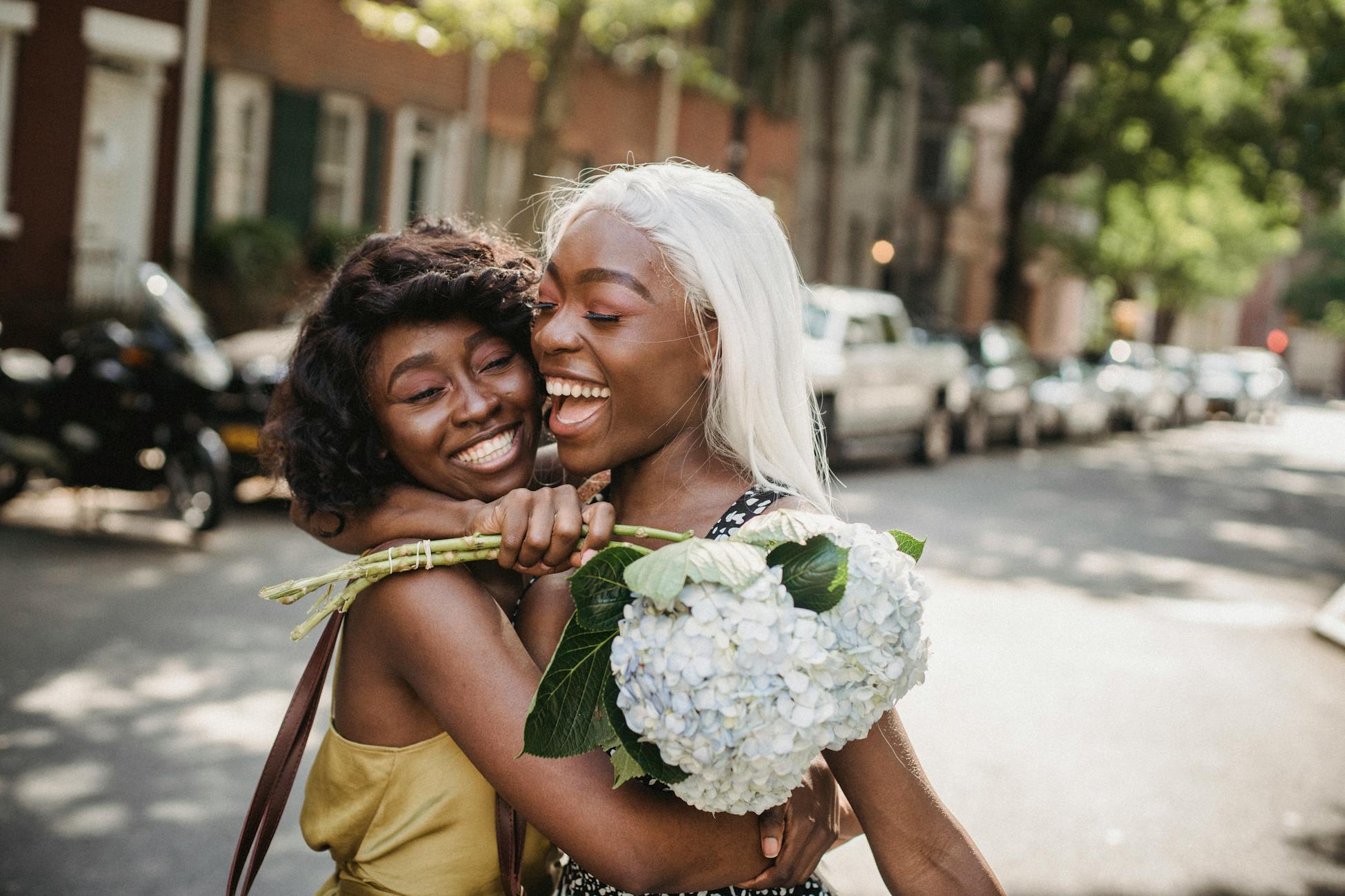 Two smiling women embrace on a sunny urban street, sharing joyful moments with a bouquet.