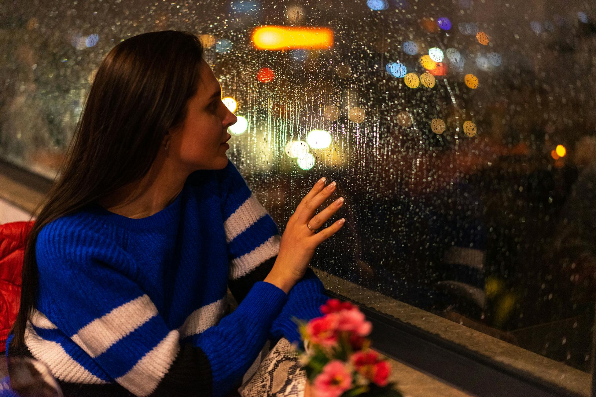 A woman in a blue sweater looks out a rainy window, reflecting city lights.