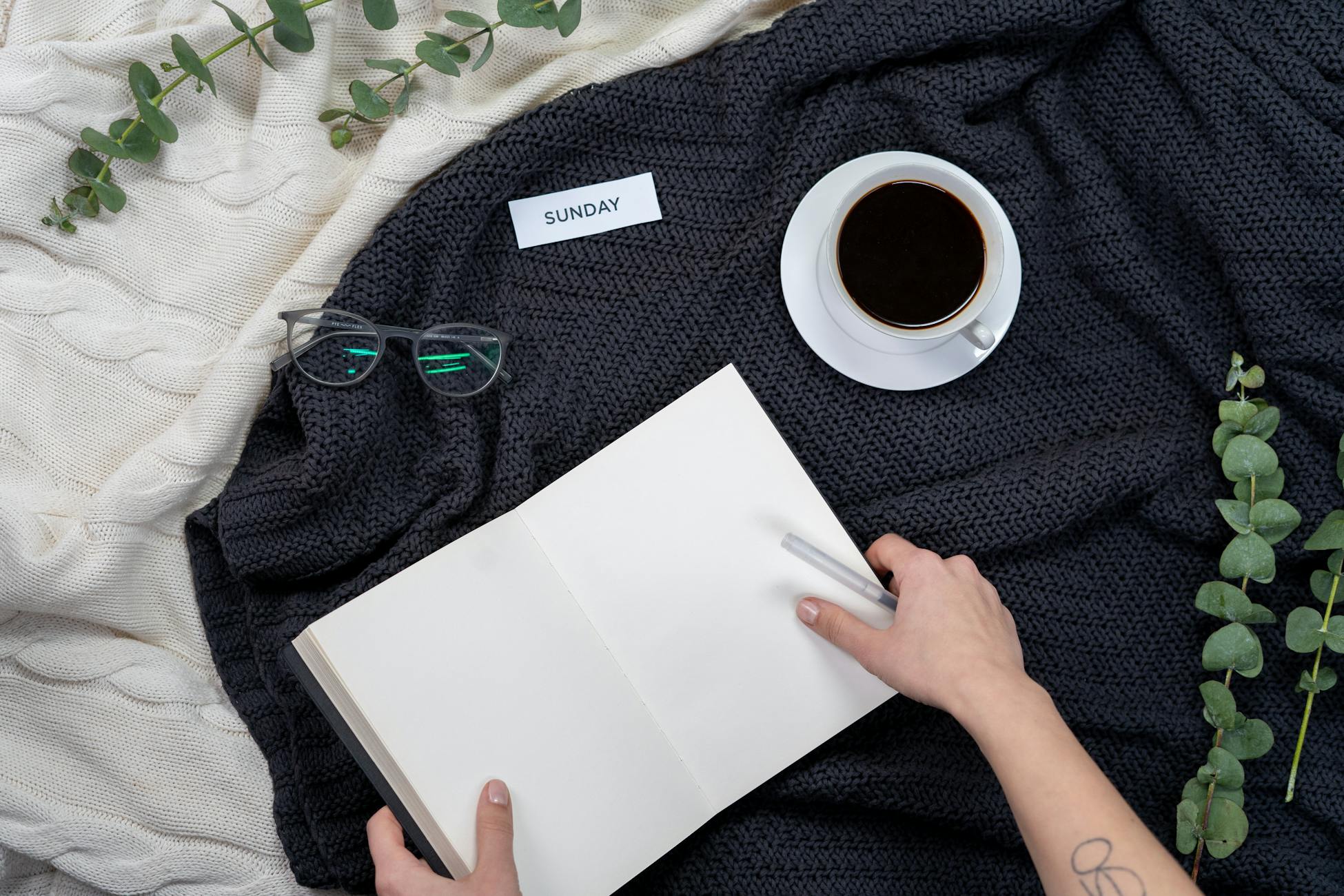 A cozy setup with black coffee, notebook, glasses, and a Sunday note on a knitted blanket.