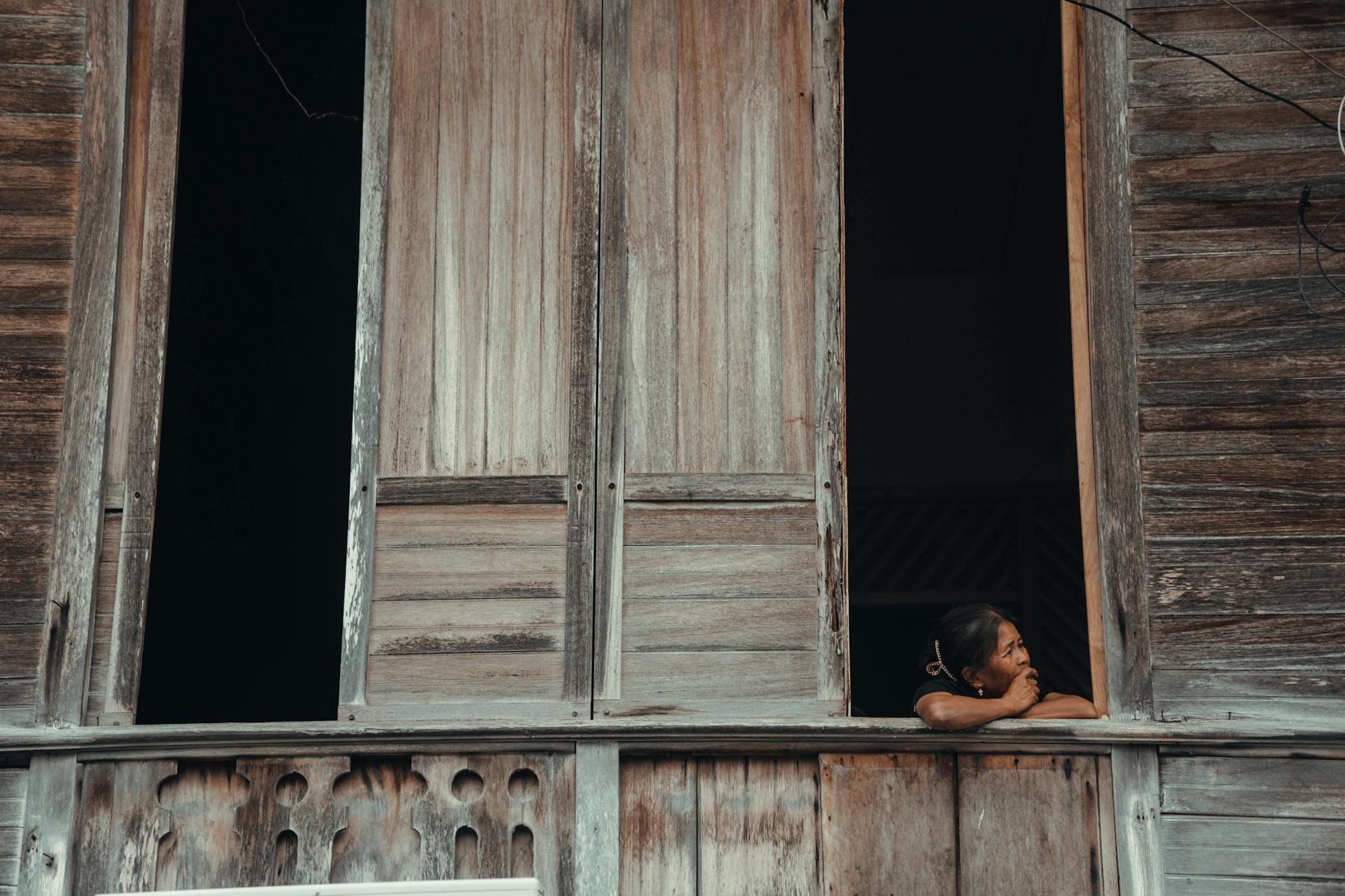 An elderly woman gazes thoughtfully from a rustic wooden window in a rural setting.