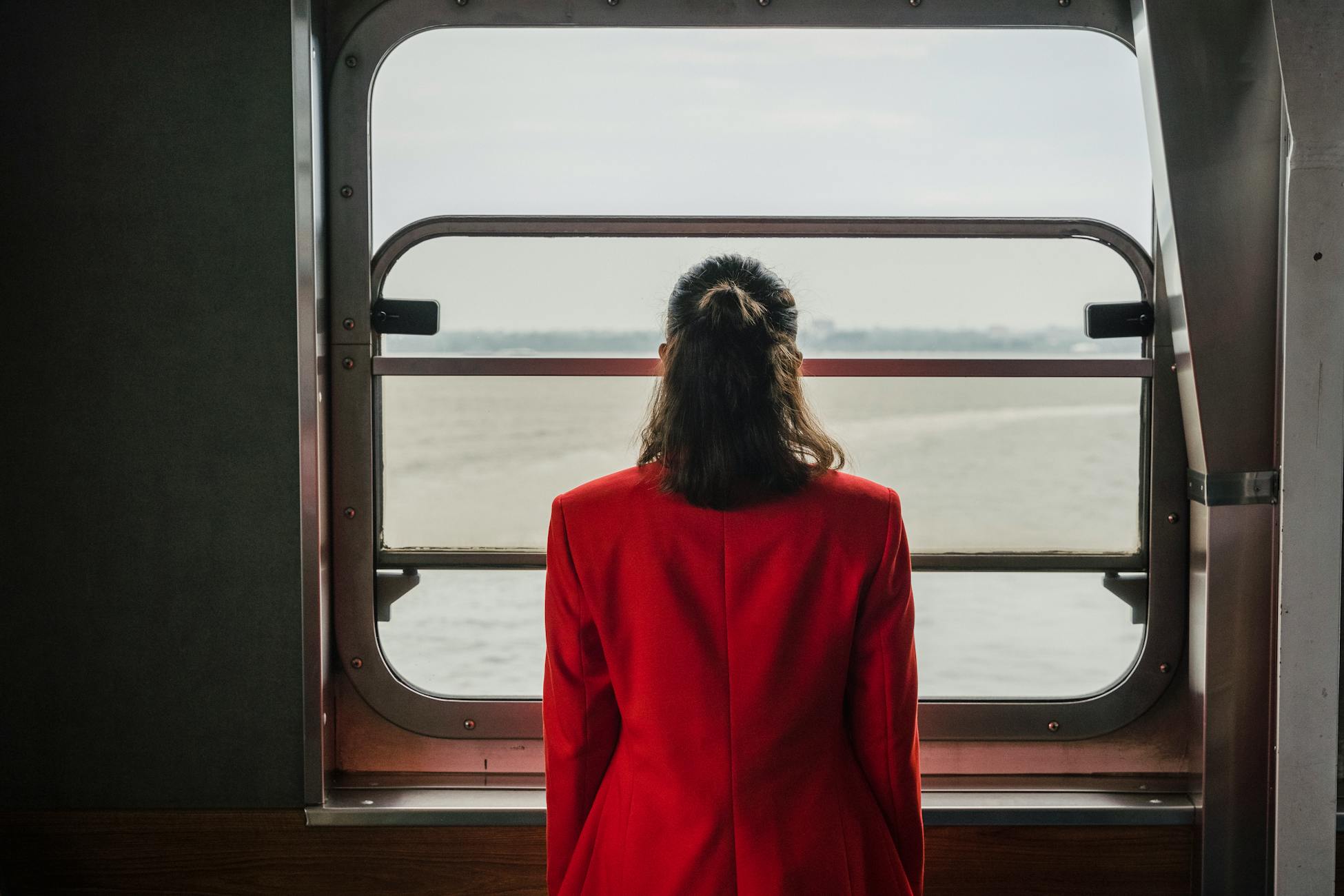 Woman in a red suit standing contemplatively by a window on a boat.