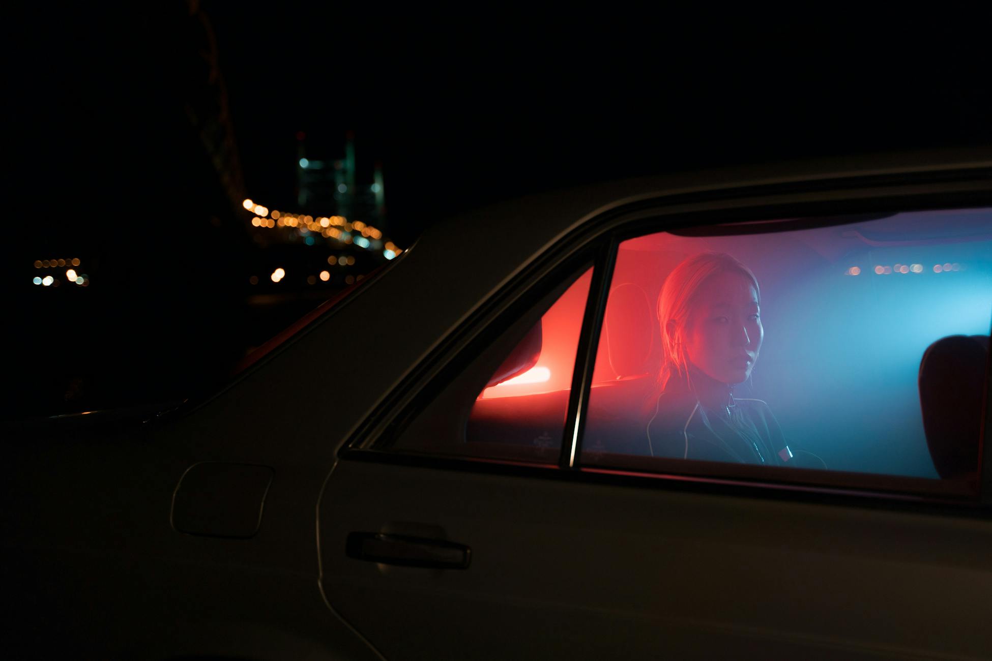 A woman sitting in a car at night with colorful lighting and city skyline in the background.