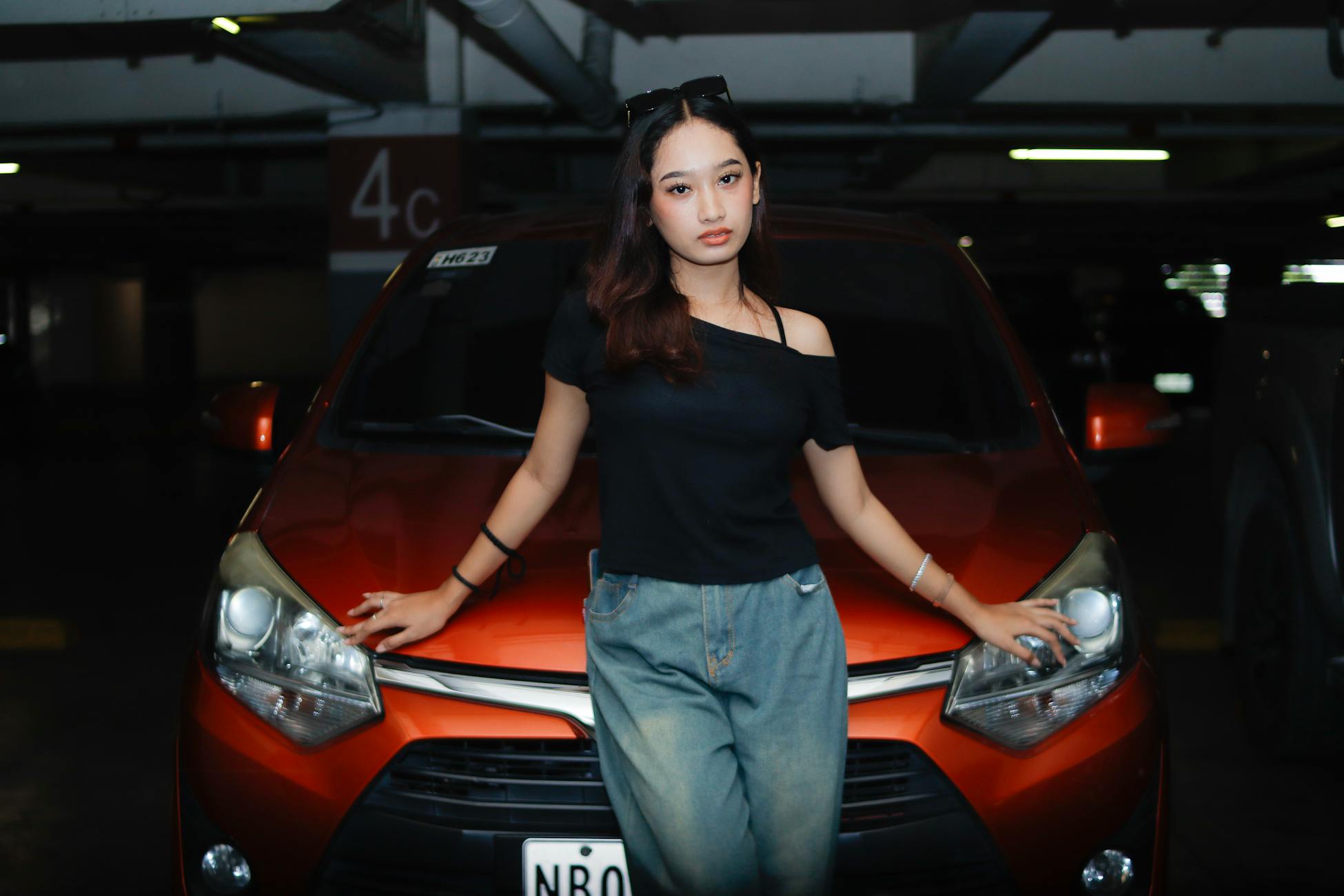 Stylish woman in a dark garage leaning on an orange car, exuding confidence.