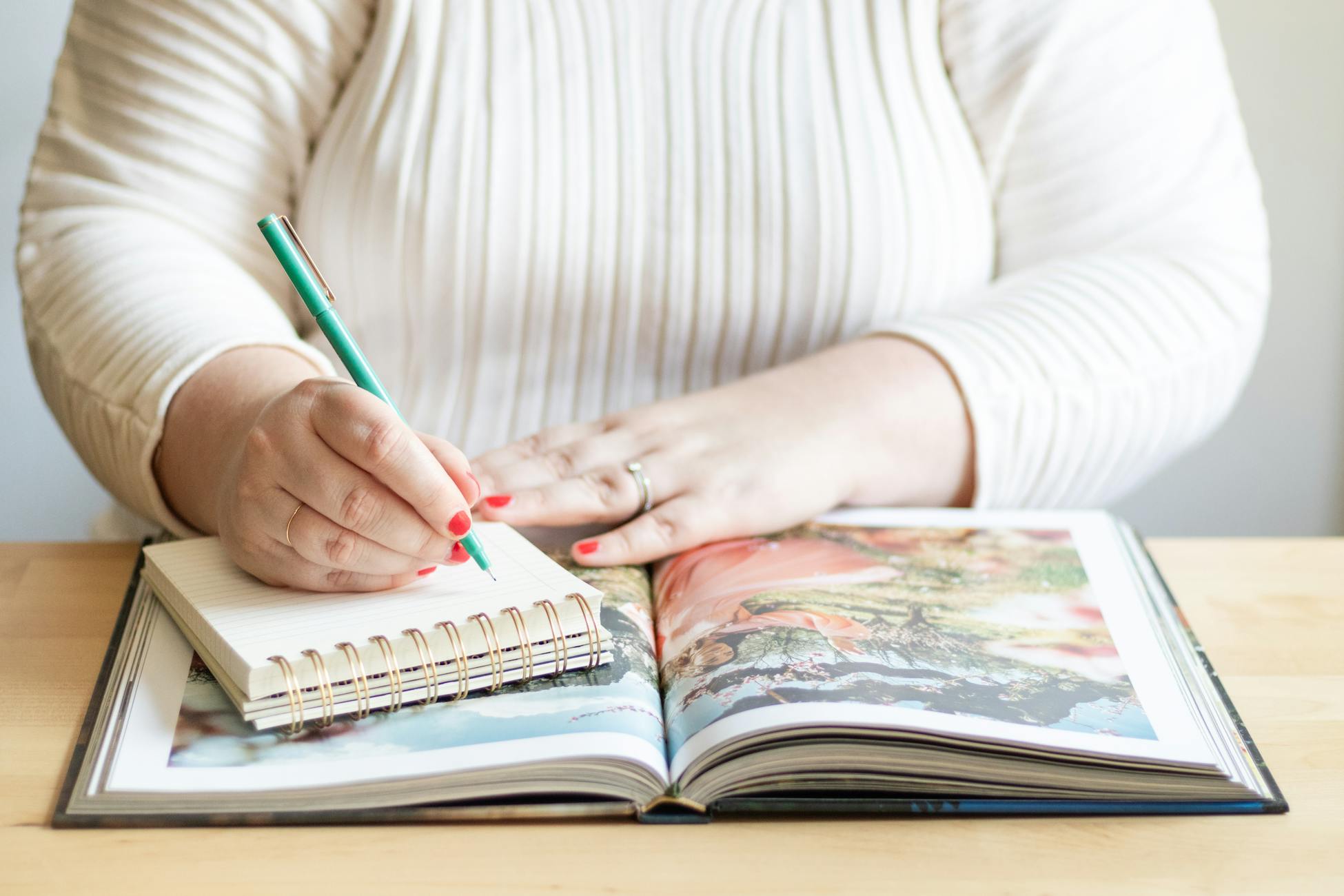 A woman writes in a notebook on a table with an open book and pen.