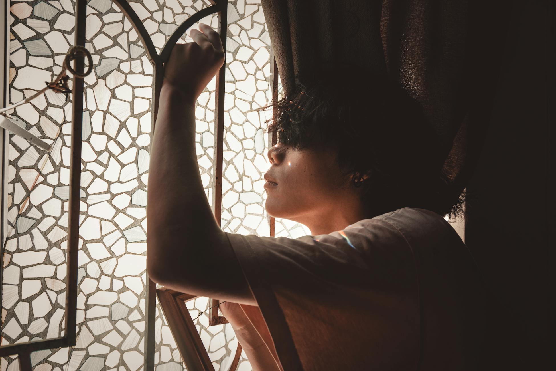 Side view of a young man posing thoughtfully by a mosaic glass window with soft light.