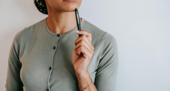 Crop anonymous ethnic female with dark hair in casual outfit touching face with pen and thoughtfully looking away against white wall