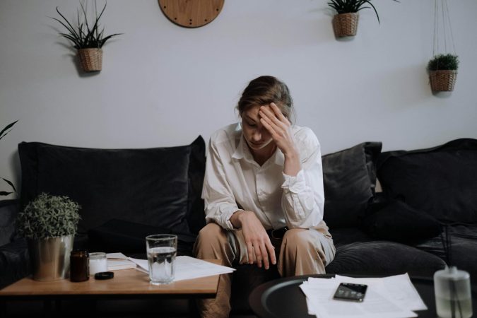 Woman experiencing stress and headache while sitting on a sofa at home, surrounded by papers.