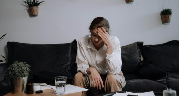 Woman experiencing stress and headache while sitting on a sofa at home, surrounded by papers.