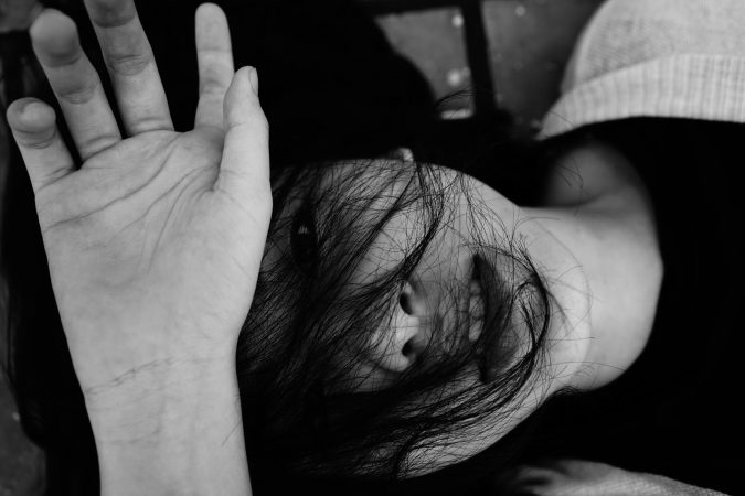Black and white top view of anonymous emotionless young lady lying on surface with dark hair and hand on face in daytime