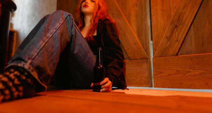 A woman sitting on the floor holding a beer bottle, contemplating indoors with warm lighting.