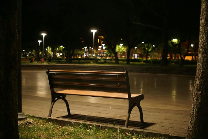 A solitary wooden bench in a quiet park illuminated by streetlights at nighttime.