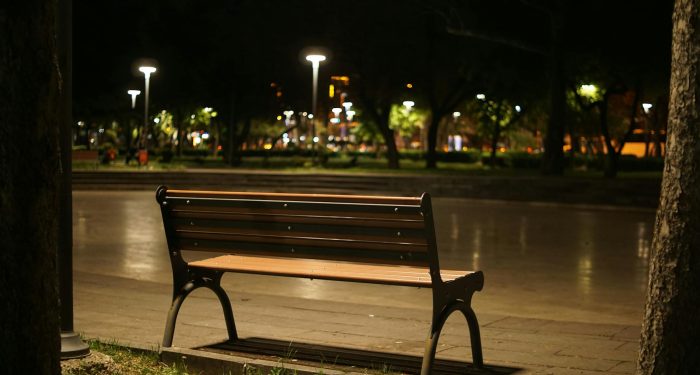 A solitary wooden bench in a quiet park illuminated by streetlights at nighttime.