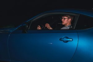 A young man in glasses driving a sleek blue car at night, showcasing modern automotive design.