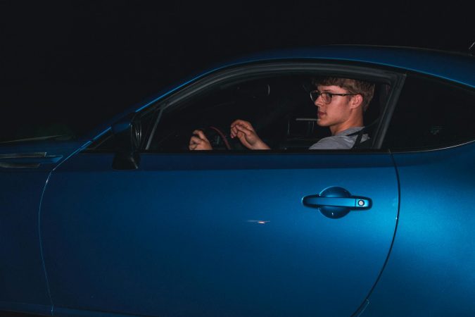 A young man in glasses driving a sleek blue car at night, showcasing modern automotive design.
