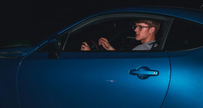 A young man in glasses driving a sleek blue car at night, showcasing modern automotive design.