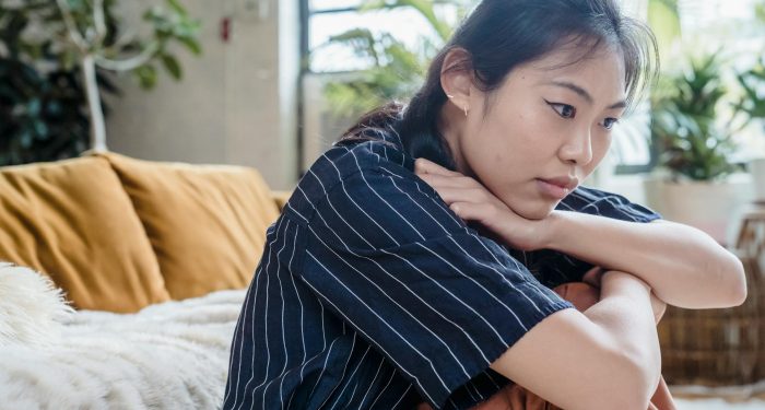 Young woman sitting pensively on a cozy couch indoors, surrounded by plants. Thoughtful and contemplative mood.
