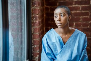Young ill bald African American female in blue patient gown looking away in window