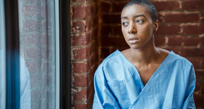 Young ill bald African American female in blue patient gown looking away in window