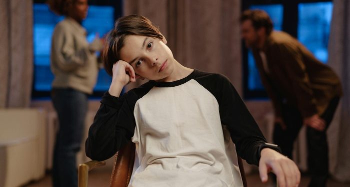A young boy in a black and white shirt looks sad as his parents argue in the background.