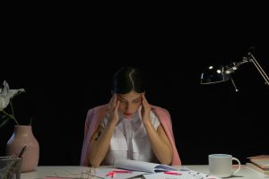A woman sitting at a desk with her hands on her head, appearing stressed and tired with office supplies around her.