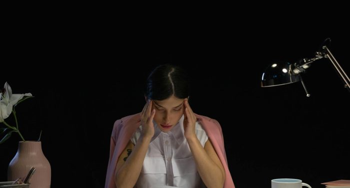 A woman sitting at a desk with her hands on her head, appearing stressed and tired with office supplies around her.