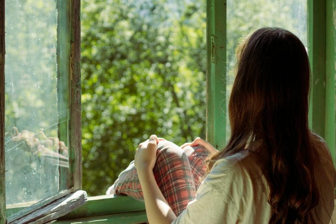 Woman sitting by an open window enjoying the view of lush greenery outside. Perfect for themes of relaxation and solitude.