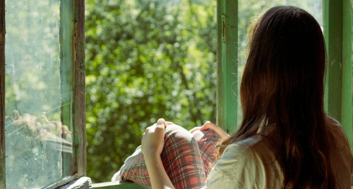 Woman sitting by an open window enjoying the view of lush greenery outside. Perfect for themes of relaxation and solitude.