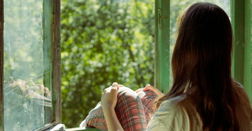 Woman sitting by an open window enjoying the view of lush greenery outside. Perfect for themes of relaxation and solitude.