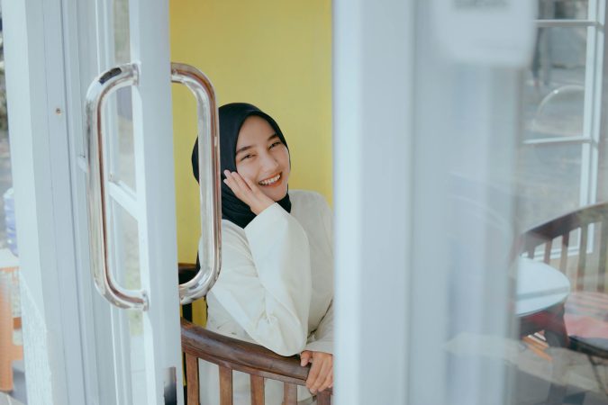 A cheerful woman in a hijab sits inside a sunlit room, smiling warmly.