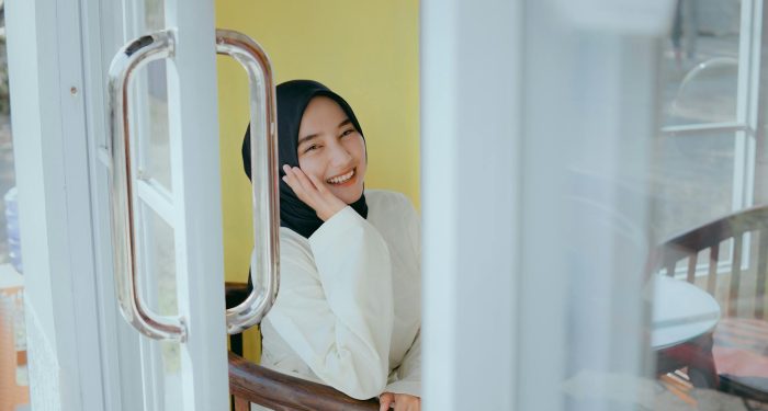 A cheerful woman in a hijab sits inside a sunlit room, smiling warmly.