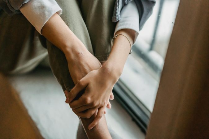 High angle of lonely crop anonymous female with bracelet standing with crossed legs and hands near window