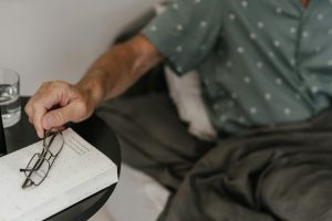 Senior adult relaxing in bed, reaching for glasses and book, creating a cozy reading moment.