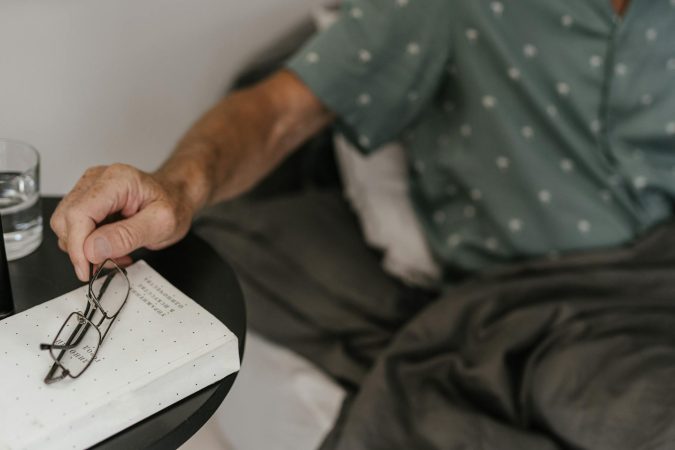 Senior adult relaxing in bed, reaching for glasses and book, creating a cozy reading moment.