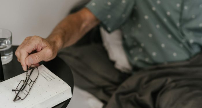Senior adult relaxing in bed, reaching for glasses and book, creating a cozy reading moment.