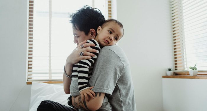 A heartfelt embrace between a parent with short hair and a child on a bed, creating a warm family scene.