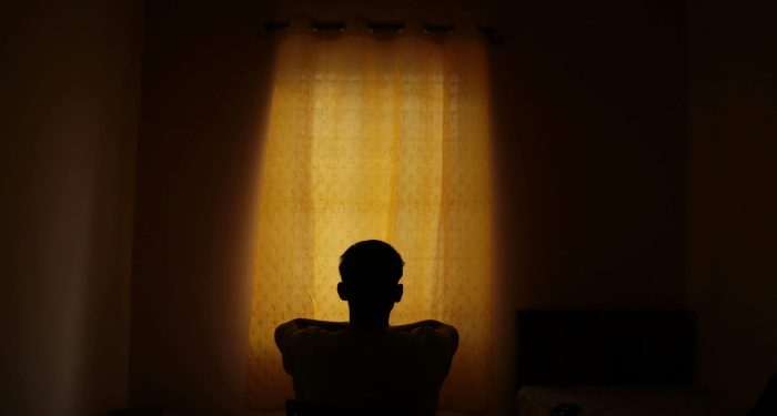 A backlit silhouette of a person sitting alone indoors, looking at a window with light filtered by curtains.