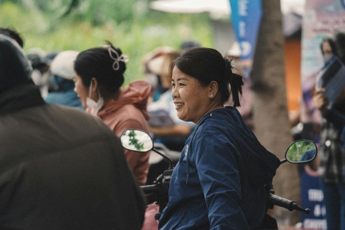 Woman in blue jacket smiles while sitting on a motorbike in a bustling outdoor setting.