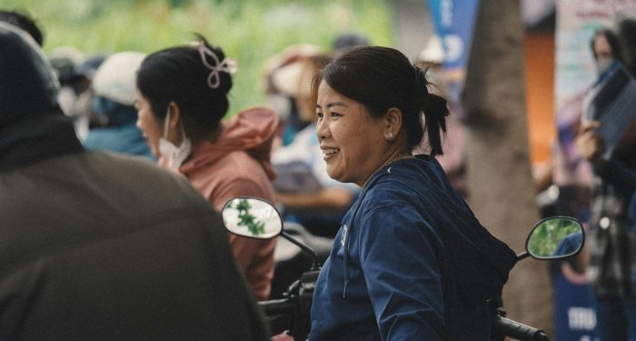 Woman in blue jacket smiles while sitting on a motorbike in a bustling outdoor setting.