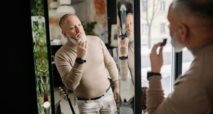 Middle-aged man applying beard care, examining reflection indoors, embracing modern grooming.
