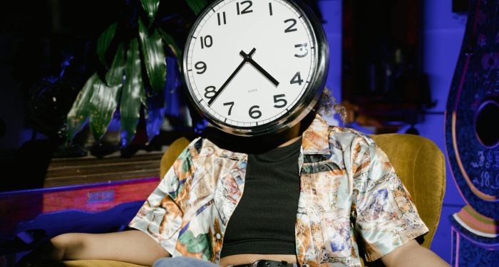 A surreal image of a person sitting in an armchair with a clock as a face, evoking themes of time and identity.