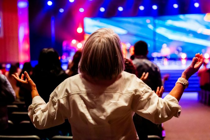 A vibrant indoor worship service with people raising hands in praise and colorful stage lighting.