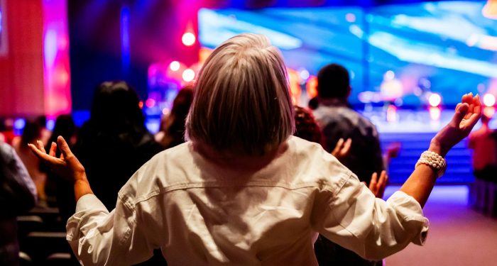 A vibrant indoor worship service with people raising hands in praise and colorful stage lighting.