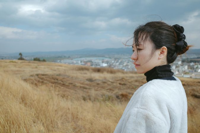 A thoughtful woman stands in a windswept field overlooking a town under cloudy skies.