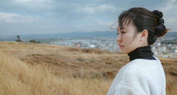 A thoughtful woman stands in a windswept field overlooking a town under cloudy skies.