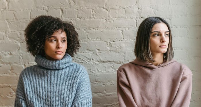 Sad African American woman with female friend in casual clothes looking away while sitting in light room during quarrel at home