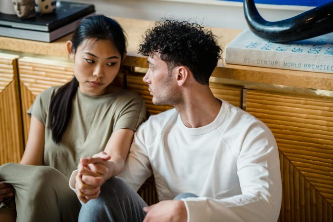 A couple sitting together indoors, holding hands with thoughtful expressions.