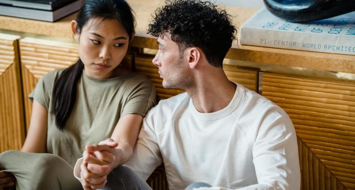 A couple sitting together indoors, holding hands with thoughtful expressions.