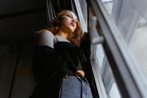 A woman gazes out of a window, deep in thought in a softly lit room.